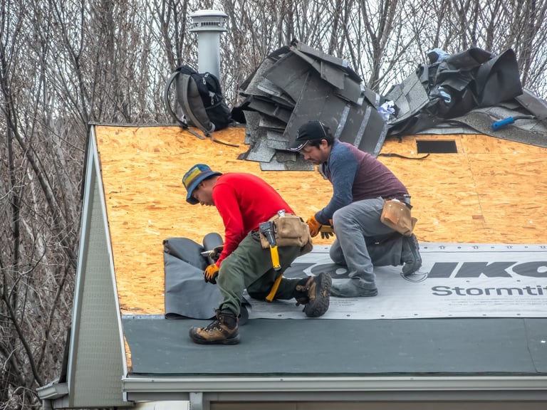 Two construction workers are installing black underlayment on a plywood roof, preparing for shingles
